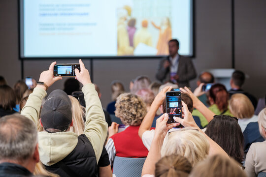 Crowd of attendees in conference hall capturing moment with phones while speaker presents on stage, energetic learning atmosphere and professional setting