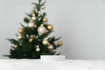 Christmas holiday background and a round miniature podium. White snow in the foreground with copy space and a blurred minimalist Christmas tree and golden ornaments.