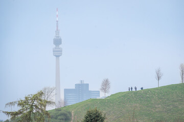 Obraz premium Urban Dortmund landscape with communication tower and silhouettes of people on grassy hill under soft blue sky.