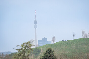 Fototapeta premium Distant city tower rising behind a green hill with bare trees and walkers enjoying a calm day outdoors.