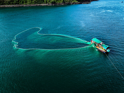 Aerial view of a fishing boat casting a wide net in the turquoise waters, contrasting with the dark green of the nearby shoreline, An Ninh Dong, Phu Yen, Vietnam.