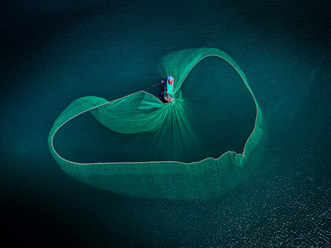 Aerial view of a fishing boat casting a wide, circular net that spreads across the dark blue waters, creating a mesmerizing dance of light and shadow, An Ninh Dong, Phu Yen, Vietnam.