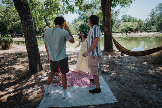 A cheerful outdoor picnic scene by a pond, featuring two men, a woman in a yellow dress, a photographer, and a hanging hammock under leafy trees. Relaxed, casual, sunlit moment.