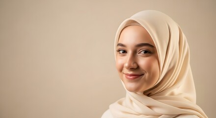 A close-up portrait of a young woman with a friendly smile wearing a beige hijab against a neutral background