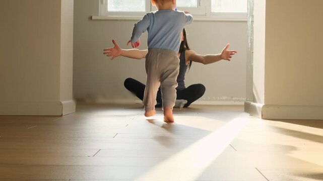 a child toddler boy with bare legs runs across the floor to his mother against a sunny window, hugging mom happy family first steps towards his family indoor