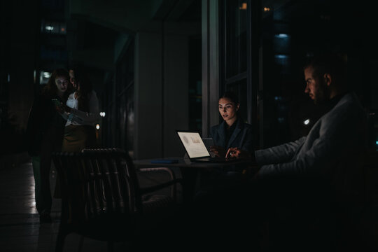 A focused woman sits at a laptop in a dim, urban setting while colleagues chat nearby, conveying professional collaboration and late-hour teamwork in a city environment.