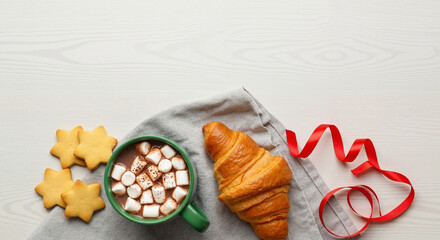Green mug filled with hot chocolate and marshmallows beside star shaped cookies and croissant on wooden table with red ribbon. Cozy winter breakfast concept for new year holiday celebrations.