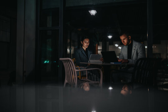 Two professionals sit at a dimly lit cafe table, focused on a laptop and tablet. The scene conveys collaboration, remote work, and evening business discussions in a modern, intimate setting.