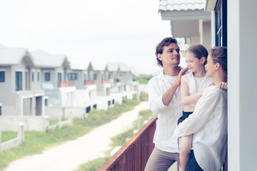 A joyful family of three enjoying a peaceful moment together on a balcony. The father, mother, and their young daughter smile and look out over a calm residential neighborhood. Perfect for themes of f