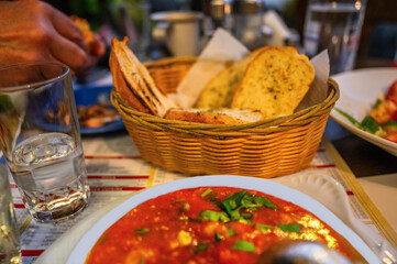 Bread in bowl, portion of shrimp saganaki, glass of water, part of hand with meal, dining at restaurant.