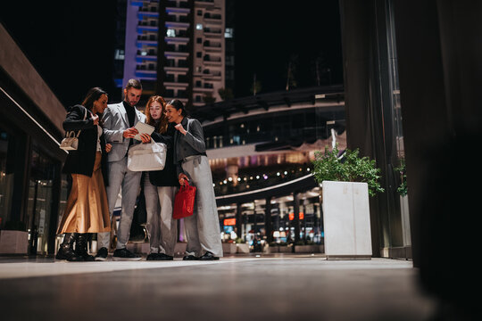 A diverse group of young professionals gathers in a modern urban plaza at night, examining a tablet together as they discuss ideas, amid shopping bags and bright city lights.