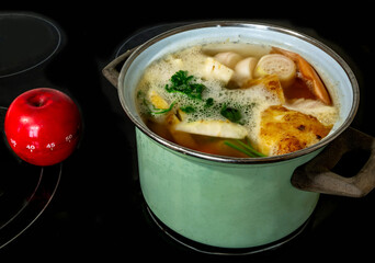 Green pot with cooking water and sliced vegetable, red apple timer on black ceramic hob.