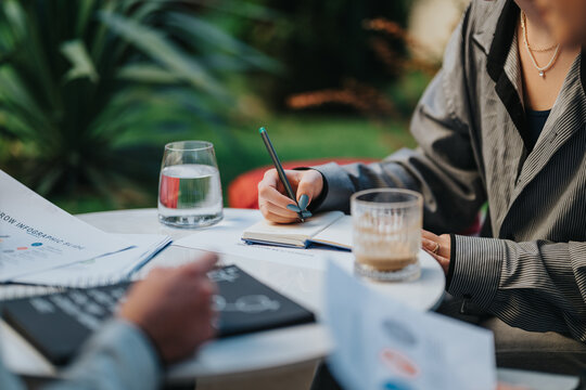 A professional outdoor setting shows a person taking notes at a round table with documents, a notebook, pen, and coffee. The scene conveys collaboration, planning, and productive work.