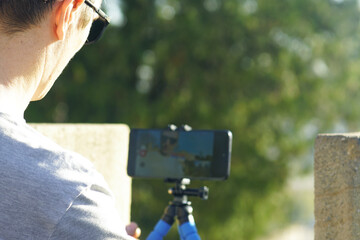 Person monitoring smartphone screen while recording video on tripod outdoors. Concept of live streaming setup, travel vlog production, self-documentation, content creation, and influencer filming.