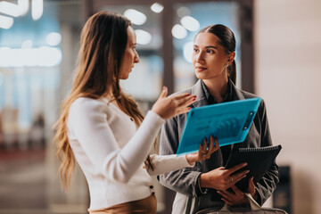 Two professional women in a modern office exchange ideas, with a blue tablet and notebook in hand. The scene captures collaboration, planning, and confident business communication.