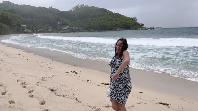 Carefree woman in black and white zebra print strapless dress dancing walks along sandy beach, embracing freedom as ocean waves roll in behind green hills.