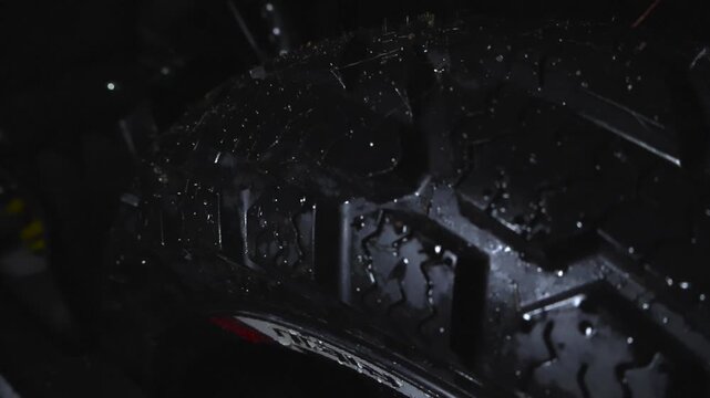 Detailed view of a wet and rain drops covered new and pristine motorcycle black tire wheel at night or evening time with bokeh background and shallow depth of field.