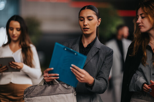 Professional women in business attire walk through a contemporary lobby, examining a blue clipboard and tablet. The scene conveys focus, collaboration, and momentum in a dynamic work environment.