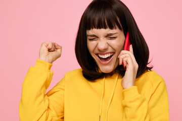 A cheerful woman in a bright yellow hoodie chats on a red phone, smiling with joy and energy as she celebrates a personal moment against a pink backdrop.