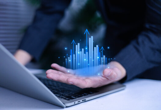 Businessman showing digital bar graph rising above laptop with glowing arrows. Concept of business growth, financial success, data analytics, and digital technology innovation.