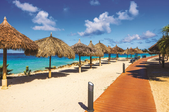 Beautiful view of wooden walkway and thatched palm leaf umbrellas on hotel beach located on Eagle Beach Caribbean Sea. Aruba.
