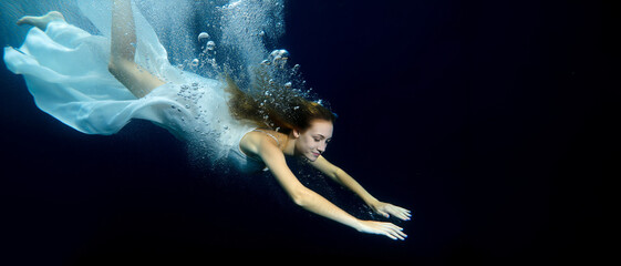 A beautiful young woman in a flowing white dress gracefully swims underwater amidst shimmering bubbles. Her serene expression and elegant movement evoke a sense of freedom and weightlessness in the de
