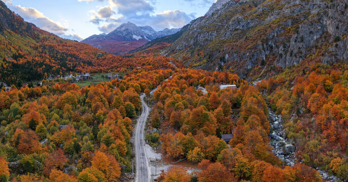 Panoramic aerial view of Valbona Valley road leading through autumn forest toward the Albanian Alps and distant Jezerca Peak.