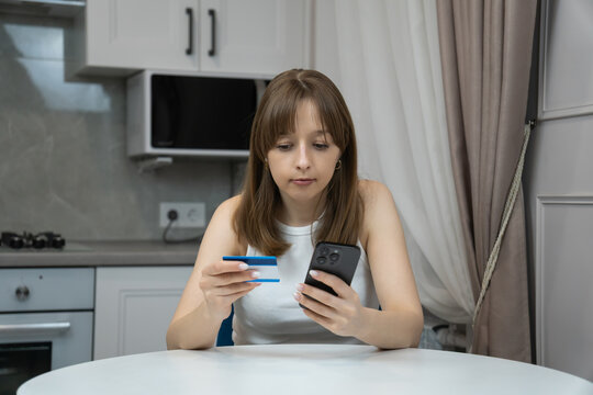 Focused young woman is using smartphone and credit card, making secure online payment or purchasing goods and services in modern kitchen at home - Powered by Adobe