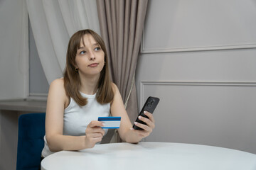 Young woman sitting at table holding credit card and smartphone, making online payment, shopping on...