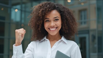Young woman smiling and raising her fist in celebration while holding a tablet outside a modern office building showing confidence and positivity