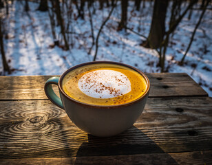 A warm cup of coffee with foam and cinnamon sits on a wooden surface in a snowy winter scene.