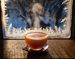 Steaming cup of tea sits on a windowsill with ice crystals forming on the glass, blurring the winter landscape outside.