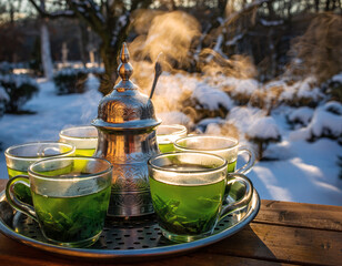 Steaming cups of green tea served from a traditional ornate metal pot on a tray in a snowy winter garden setting.
