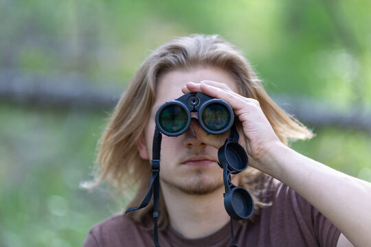 A close-up portrait of a young man with long blonde hair. He is holding a pair of binoculars with one hand, with lenses facing the camera and partially covering his face. Focus on lenses.