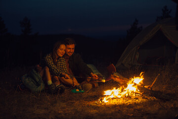 Young couple of tourists in love are sitting by the campfire against the tent, toasting marshmallows on sticks in the mountains at night.