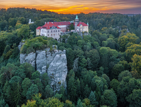 Aerial view of the majestic Hrub&aacute; Sk&aacute;la Castle perched atop rugged cliffs amidst a sea of emerald foliage under a fiery sunset, Turnov, Liberec Region, Czechia.