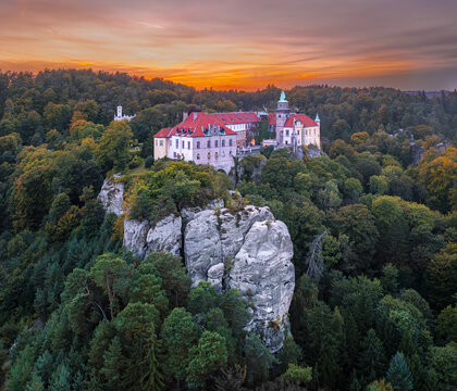 Aerial view of the majestic castle perched atop rugged cliffs amidst a sea of green foliage under a fiery sunset sky, Turnov, Liberec Region, Czechia.