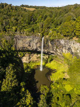 Aerial view of a slender waterfall gracefully cascading down a rugged cliff into a dark pool, embraced by vibrant green forest, Raglan, Waikato Region, New Zealand.