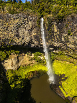 Aerial view of a powerful waterfall cascading down a rocky cliff face, plunging into a dark pool surrounded by vibrant green moss, Raglan, Waikato Region, New Zealand.