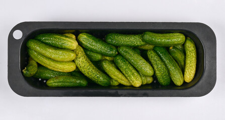 A top-down view of freshly harvested, bumpy, green pickling cucumbers (gherkins) in a black plastic strainer against a bright white background