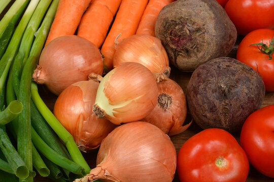 A vibrant close-up of a variety of fresh, raw vegetables, including yellow onions, orange carrots, red beets, ripe tomatoes, and bright green scallions