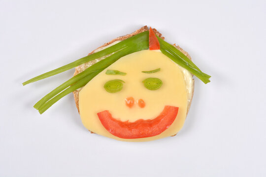 A creative, smiling face made from a slice of cheese, tomato, and green onions placed on a piece of bread, set against a plain white background