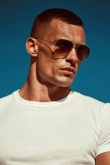 Fototapeta premium A handsome male model wearing sunglasses and a white t-shirt stands against a blue sky background, with short hair, sunlight, an upper body portrait.