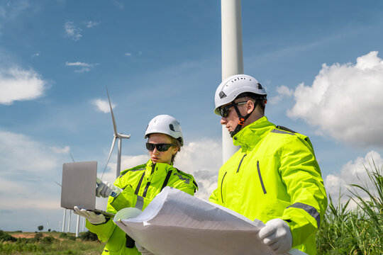 Two people wearing yellow jackets and helmets are looking at a laptop. One of them is pointing at the screen. Low angle view of two technicians (man and woman) inspecting wind turbine structure.