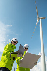 Fototapeta premium Two people are standing next to a wind turbine. One of them is holding a laptop. Low angle view of two technicians (man and woman) inspecting wind turbine structure.