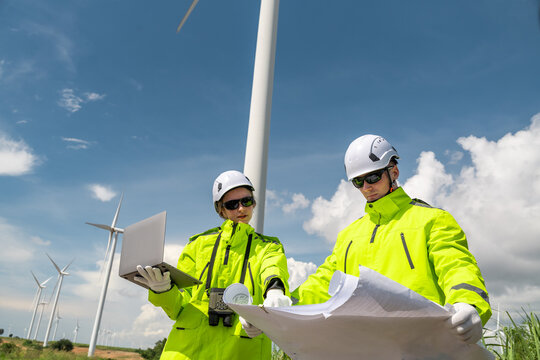 Two men are standing in front of a wind turbine. One of them is holding a laptop. Low angle view of two technicians (man and woman) inspecting wind turbine structure.