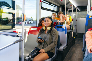 Young latina woman with a prosthetic leg smiling, holding a phone, and wearing headphones while traveling on a public bus