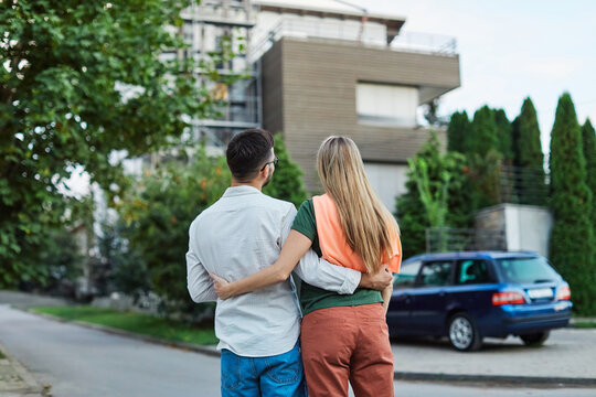 Portrait of a happy young couple standing in front of their new house, with keys in hands, smiling man and woman posing near modern home, making selfie, real estate agency, house owners, young family