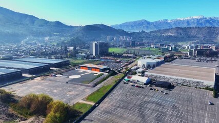 Forward drone aerial of industrial park with warehouses and parking lots, dramatic Andes mountains with snow-capped peaks backdrop, Chile - Powered by Adobe