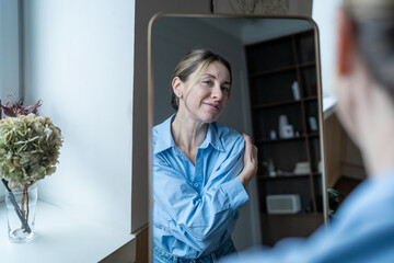 Happy mature woman embracing herself while looks on her reflection in mirror with smile, feels confidence and self-acceptance. Self-love in hug action, practicing self-care for finding inner peace. 